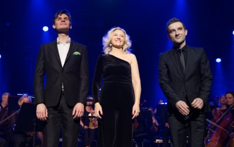 na scenie stoją obok siebie uśmiechnięci dwaj mężczyzni i między nimi kobieta z bląd włosami w tle muzycy orkiestry symfonicznej