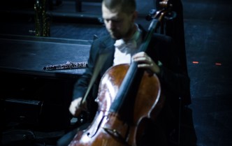 mężczyzna w ciemnym garniturze siedzi i gra na wiolonczeli zanim stoi kobieta z długimi blond włosami i gra na skrzypcach                        grająca na skrzypcach