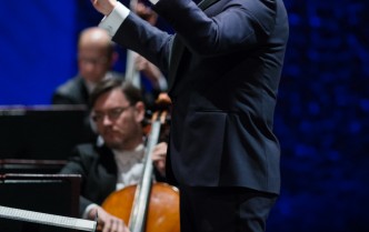 męzczyzna-dyrygent z ciemnymi włosami z bródką stoi z uniesionymi rękami w prawej dłoni trzyma batutę dyrygencką w tle lekko zamazany siedzi mężczyzna i gra na wiolonczeli