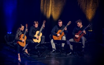 siedzący na scenie czworo muzyków kobieta i trzech mężczyzn ze skupionymi twarzami graja na gitarach