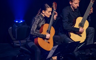 kobieta z ciemnymi związanymi w ogon włosami i mężczyzna w ciemnej koszuli siedzą na scenie grając na gitarach