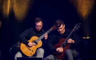 dwóch mężczyzn w ciemnych koszulach i popielatych spodniach siedzą na scenie grając na gitarach