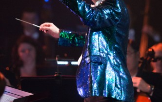 mężczyzna w świecącej niebieskiej marynarce z uniesionymi rękami i batą w prawej dłoni stoi na scenie za nim zamazani muzycy orkiestry