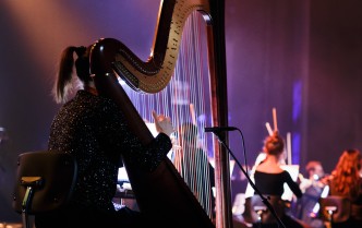na scenie sali koncertowej tył siedzącej kobiety grającej na harfie w tle też z tyłu muzycy grający na instrumentach smyczkowych