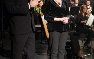 na scenie sali koncertowej stoją mężczyzna w ciemnym garniturze w prawej dłoni trzyma mikrofon w lewej bukiet czerwonych róż obok kobieta z krutkimi blond włosami z dłońmi splecionymi przed sobą w dali siedzą muzycy orkiestry