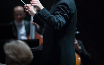 mężczyzna-dyrygent z brudką w ciemnym fraku stoi przed muzykami z uniesionymi rękoma w prawej dłoni trzyma batutę dyrygencką