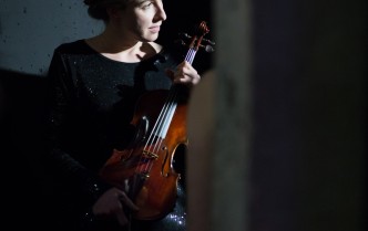 kobieta siedzi w ciemnym stroju z włosami upiętymi z tyłu głowy, patrzy w bok w dłoniach przed sobą trzyma skrzypce