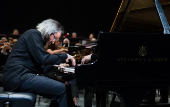 mężczyzna w okularach ze skupioną twarzą gra na fortepianie w tle muzycy orkiestry symfonicznej