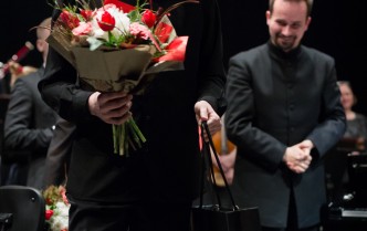 na scenie stoi mężczyzna w ciemnym stroju w okularach z zadowoloną lekko uśmiechniętą twarzą w prawej dłoni trzyma buliet z biało czerwonych kwiatów za nim po prawej stoi uśmiechnięty mężczyzna z brudką z dłońmi założonymi na siebie