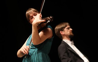 kobieta w długiej zielonej sukni grająca na skrzypcach, na drugim planie mężczyzna grający na fortepianie