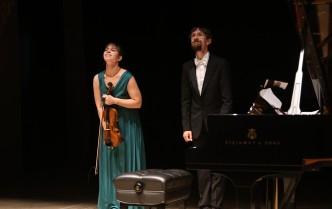kobieta w długiej zielonej sukni trzymająca skrzypce oraz mężczyzna w garniturze stojący przy fortepianie kłaniają się przed widownią