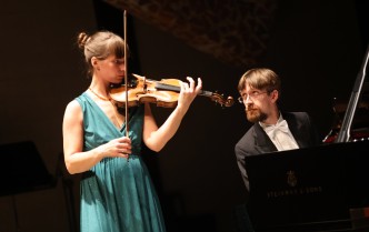 kobieta w długiej zielonej sukni grająca na skrzypcach, na drugim planie mężczyzna grający na fortepianie