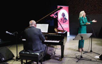 mężczyzna grający na fortepianie, obok kobieta w zielonym garniturze śpiewa z wyciągniętą do góry dłonią