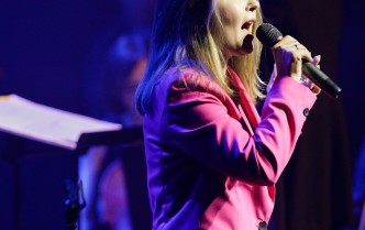 kobieta w różowym garniturze śpiewająca do mikrofonu, w tle dyrygentka