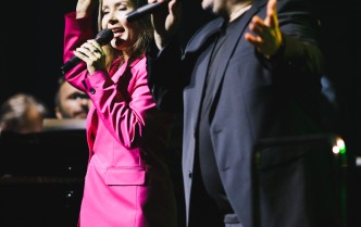 kobieta w różowym garniturze wraz z mężczyzną w ciemnym, śpiewają trzymając mikrofony w dłoniach