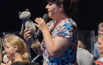 kobieta w kolorowej sukni trzymająca w dłoni mikrofon, a w drugiej pluszowego kota