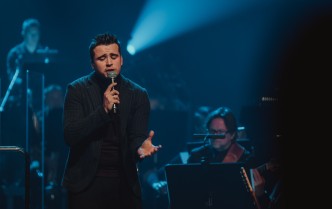 mężczyzna w ciemnym stroju na scenie z niebieskim światłem śpiewa do mikrofonu