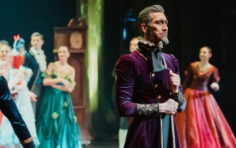 mężczyzna przebrany w ciemnofioletowy płaszcz z postarzającą charakteryzacją twarzy patrzący przed siebie, w tle inne postacie baletu