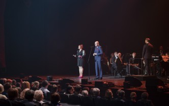 kobieta w garniturowej sukni z mężczyzną w garniturze stoją na scenie przemawiając