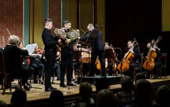 dwóch mężczyzn grających na waltorniach, w tle siedzi orkiestra