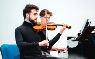 zbliżenie na skrzypka grającego na instrumencie, ukazany od pasa w górę, za nim mężczyzna grający na fortepianie