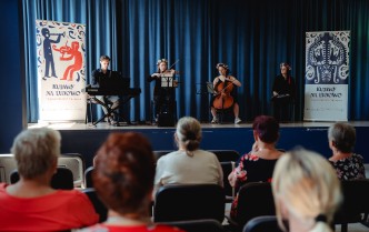 grupa muzyków grających na scenie, obok których siedzi kobieta prowadząca koncert, w dolnej części fotografii widać publiczność oglądającą koncert