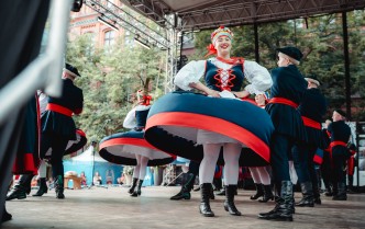 kobieta tańcząca z mężczyzną w strojach kujawskich, za nimi inne pary taneczne, kobieta ma suknię uniesioną do góry przez taniec