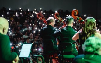 troje muzyków grających na kontrabasie ukazanych od tyłu, za nimi zaciemniona widownia z publicznością z drobnymi światełkami