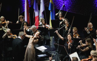 kobieta grająca na skrzypcach, na drugim planie siedzą osoby grające na skrzypcach