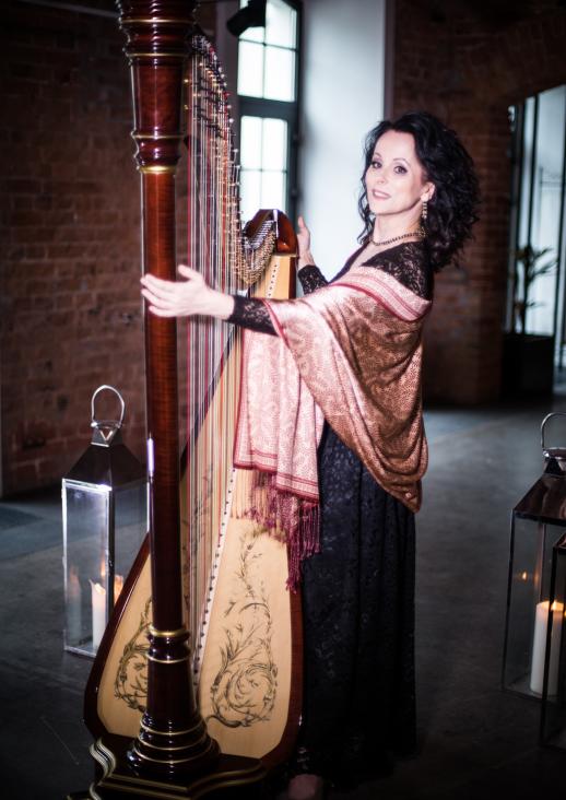 a woman with dark curly hair with a scarf over her shoulders standing with a harp