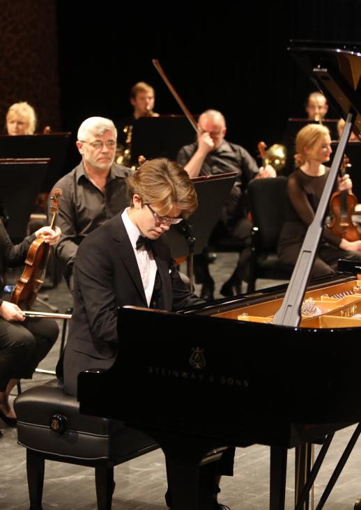 a young man with blonde hair playing the piano, behind him are women and a man with a violin in his hands
