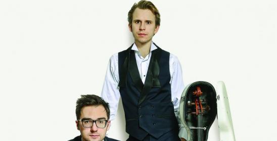 młody siedzący mężczyzna i młody stojący mężczyzna, obok którego stoi wiolonczela w otwartym futerale