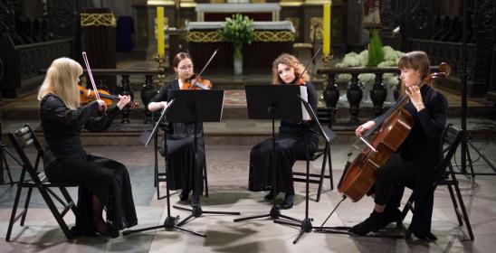 four young women in dark dresses playing stringed instruments sitting in front of music stands