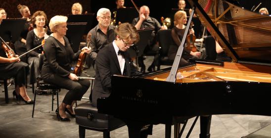 młody mężczyzna o blondowych włosach grający na fortepianie, za nim siedzą kobiety i mężczyzna ze skrzypcami w rękach