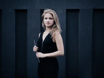 blonde young woman standing against a dark wall holding a baton in her hands