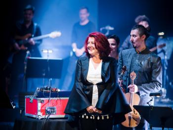 Beethoven Electric 2D solistka na pierwszym planie, w tle muzycy trzymający instrumenty muzyczce, Solistak iubrana z czarna połyskujaca marynarkke i białą koszulę, uśmiechająca się, twarz skierowana ku prawej stronie