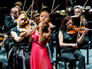Uczestniczka konkursu skrzypcowego, ubrana w czerwona długa plisowana suknię, w tle muzycy, sekcja skrzypiec. Kobieta gra na skrzypcach, ujęcie w centrum.