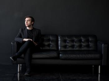 A young man with dark hair in a black shirt sitting on a black couch