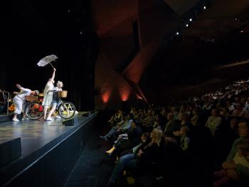 na scenie trójka artystów, jeden na rowerze, drugi trzymający parasol, trzeci ciągnący rower do tyłu. Z lewej strony widać  publiczność 
