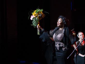 Festiwal Nova Muzyka i Architektura ciemnoskóra artystka ubrana w ciemną długa suknię, trzymająca bukiet kwiatów koloru żółtego. Scena , reflektor skierowany na artystkę.