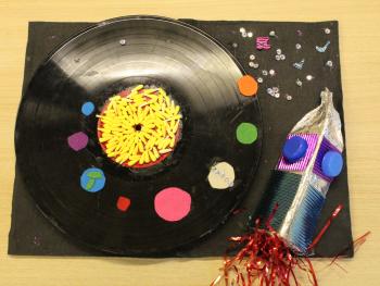 Praca plastyczna dziecka przedstawiająca artystyczną interpretację kosmosu, gdzie planetę tworzy płyta winylowa na której znajdują się kolorowe kulki a obok rakieta kosmiczna. 