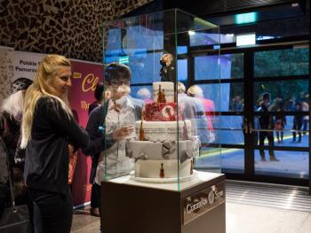 Festiwal Nova Muzyka i Architektura imitacja tortu piętrowego, na podeście za szybą, spogladający na niego Melomanii, tort zawiera elementy muzyczne