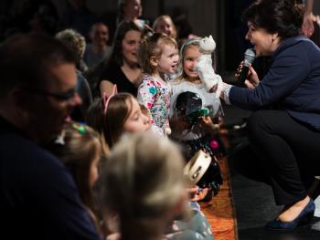 Dni Moniuszkowskie - koncert dla dzieci - Pies i kotek koncert dla dzieci. lewa strona dzieci wraz z rodzicami siedzący na pomarańczowym dywanie. Dzieci trzymają instrumenty muzyczne, po prawej stronie prowadząca koncert, ciemnowłosa kobieta ubrana w granatowy garnitur, na lewek ręce ma założona pacynkę białego kotka, w prawej trzyma mikrofon