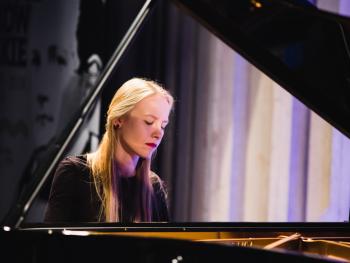 Dni Moniuszkowskie - Recital fortepianowy młoda pianistka grająca na fortepianie, zbliżenie na twarz, w tle rollup Dni Moniuszkowskich, przedstawiający graficzną postać Moniuszki