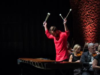 Festiwal Siły Natury - Moc ognia młody mężczyzna grający na marimbie, ubrany w czerwona koszulę, ręce skierowane ku górze, w tle ściana sali koncertowej CKK Jordanki - tzw. pikado.