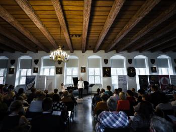 Festiwal Nova Muzyka i Architektura Widok na scenę w Sali Ratusza Staromiejskiego, w tyle okna i rollpy, centralnie widoczne przejście pomiędzy krzesłami ustawionymi na widowni. Widownia pełna, widoczni atyści