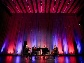 ujęcie Kwartetu (dwie kobiety i dwóch mężczyzn) podczas koncertu online odbywającego się w Sali Prób. Kolorowe światła (czerwono-fioletowe) padają  na ściany oraz sufit sali.  