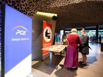 podświetlany rollup PGE Energia Ciepła SA na foyer, w tle Melomani kierujący się w stronę sali koncertowej, kobiety biorące repertuary, eksponowane na stoliku nieopodal 