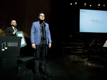 Mężczyzna przemawiający do mikrofonu na scenie, w tle logotyp Weyna Invest oraz mężczyzna trzymający oprawione w czarnej ramie podziękowania