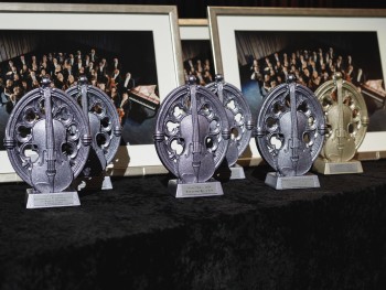 sześć statuetek w kształcie skrzypiec wkomponowanych w okrągłe gotyckie rozety. W tle oprawione w złote ramy zdjęcie orkiestry.